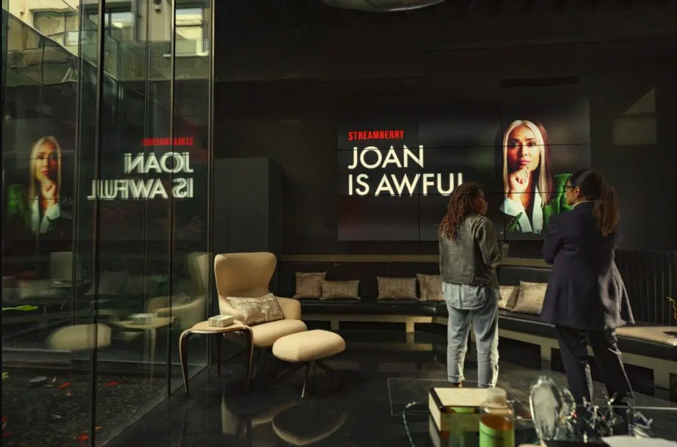 Scena z serialu "Black mirror" z odcinka "Joan jest okropna". Dwie kobiety stoją w eleganckim, ciemnym pokoju i patrzą na telewizor. Odtwarzają serial "Joan is awful" na platformie "Streamberry".