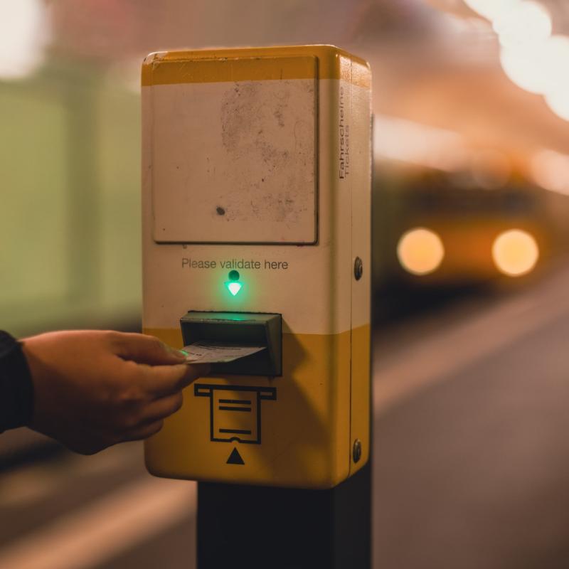 Osoba wkłada bilet do elektronicznego kasownika. W tle nadjeżdżający pociąg metra